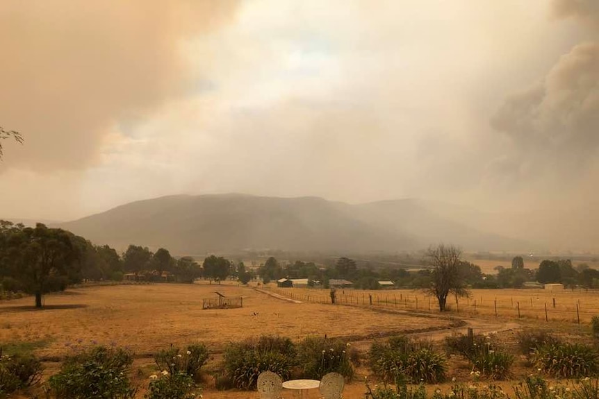 Brown-grey smoke billows over hills and the dry grass of a property.