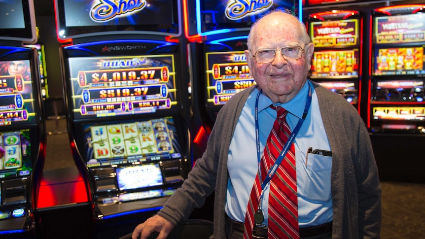 Len Ainsworth smiles as he stands in front of a row of flashing Ainsworth Quad Shot pokie machines.