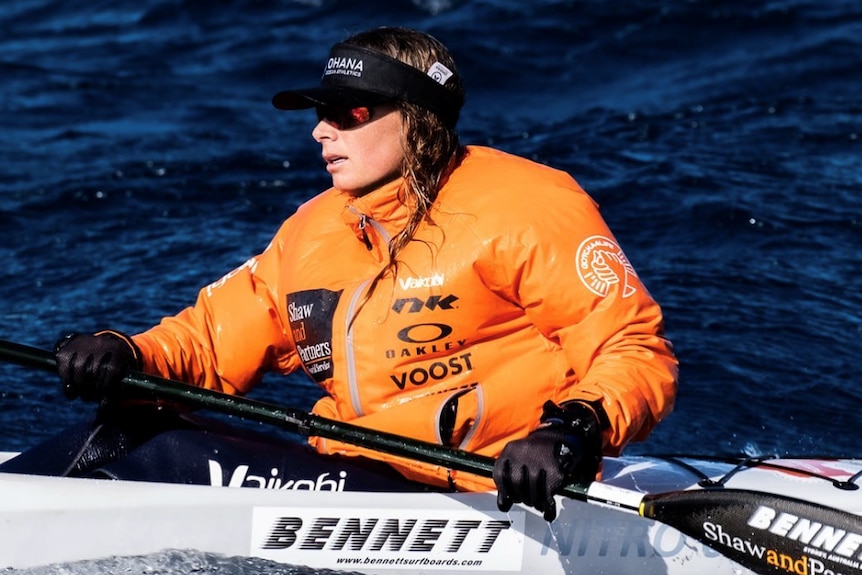 Woman on surf ski holding a paddle and wearing an orange water jacket.
