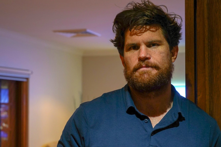 A man in a blue shirt leaning on a door frame inside a house