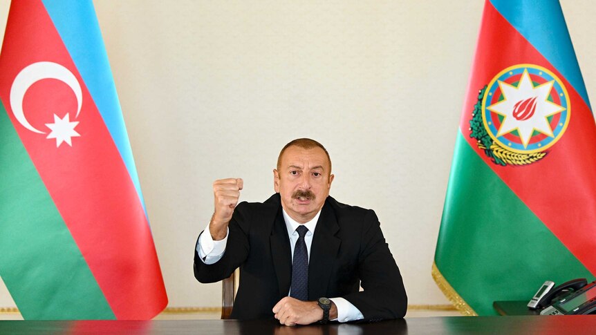 Azerbaijani President Ilham Aliyev, with short hair and a moustache, gestures in a TV address between two flags.