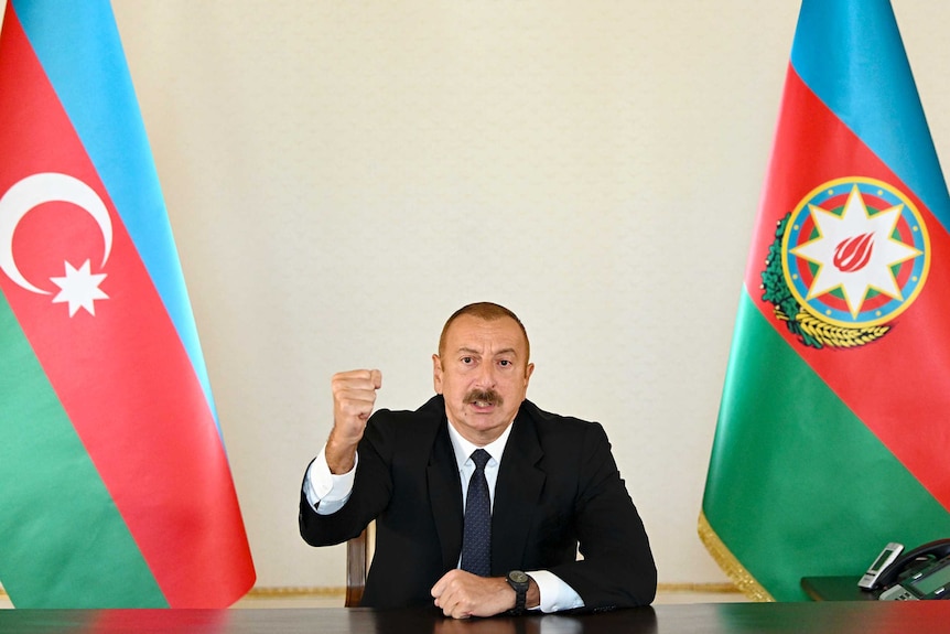 Azerbaijani President Ilham Aliyev, with short hair and a moustache, gestures in a TV address between two flags.