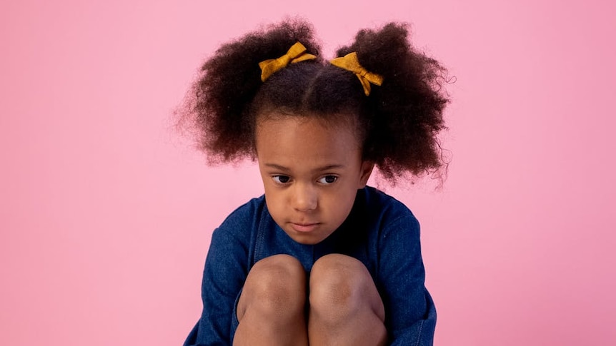 A young girl sits with her arms wrapped around her legs. She has worried expression on her face.