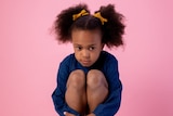 A young girl sits with her arms wrapped around her legs. She has worried expression on her face.