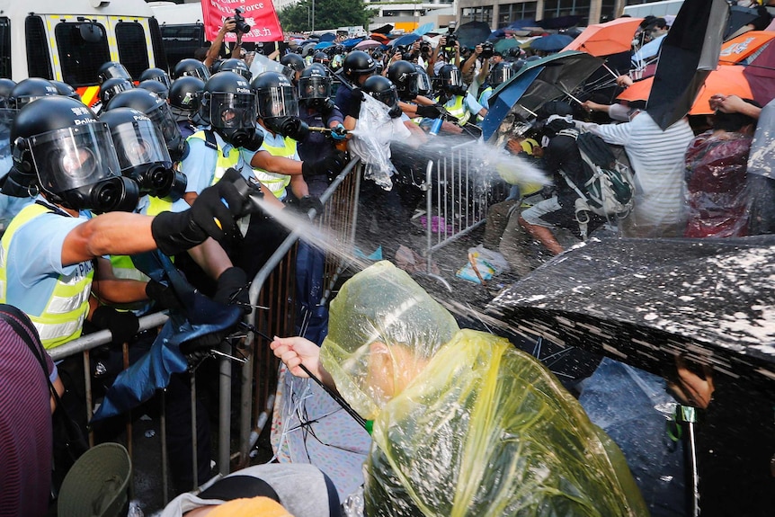 Police on one side spray protesters holding umbrellas with pepper spray.