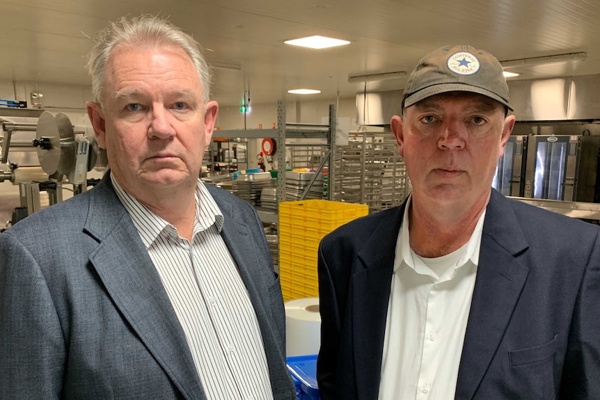 Two middle-aged men, one wearing a cap, stand in a kitchen looking blankly at the camera.