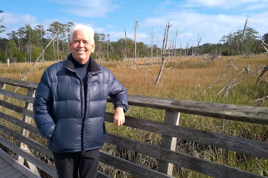 Elderly man smiling, wearing a navy puffer jacket and leaning against a wooden fence outdoors.