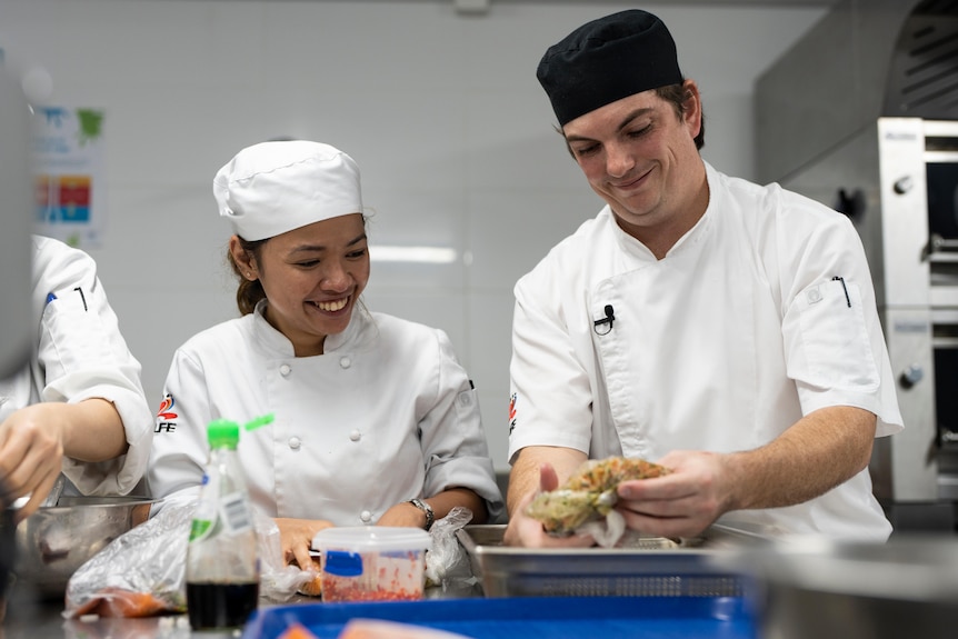 A chef in a black cap instructs a smiling trainee in a commercial kitchen.