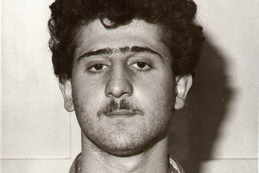 A young man with dark curly hair and a moustache poses for a mugshot.