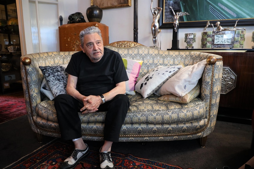 John Playfoot sits on a half circle couch covered in pillows, he leans forward and rests his elbows on his knees