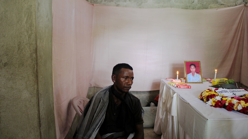 You view a man in the centre of the frame sitting next to a coffin draped in fabric, flowers and candles. 