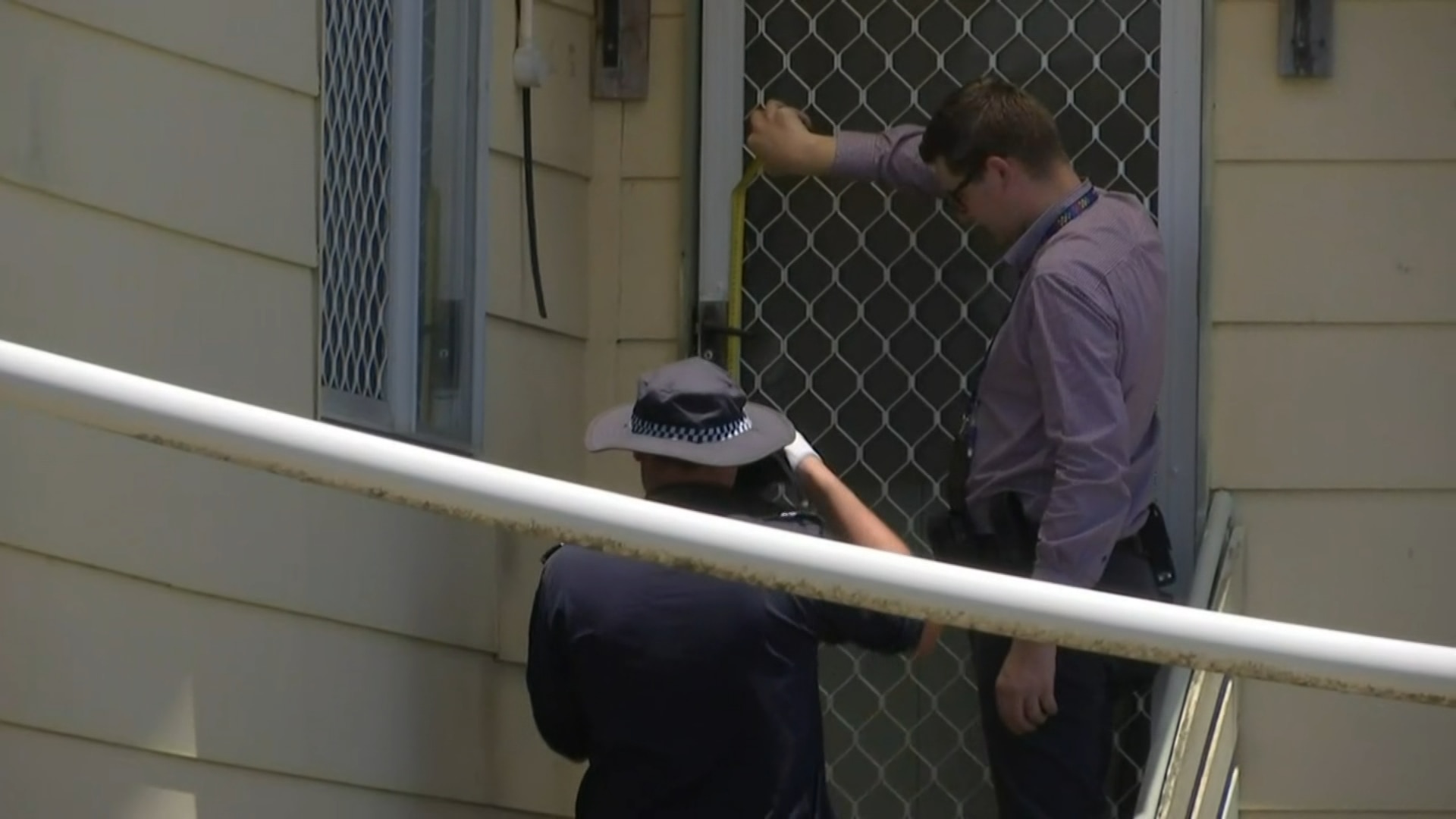 Dos hombres están parados en los escalones superiores de la entrada de una casa de dos pisos, sosteniendo una cámara y una cinta métrica. 