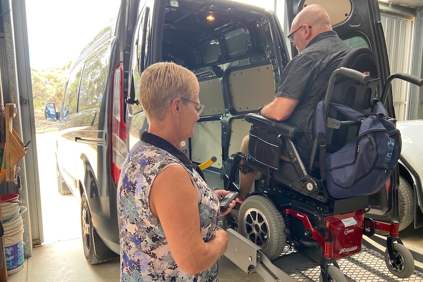 A man in a wheelchair on a lifter at the back of a van with a woman standing nearby