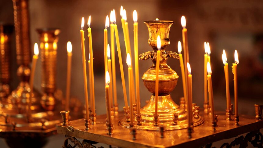 Ornate candle stick holder with many long, thin candles burning. Orange glow from candles (in a church but background blurred).