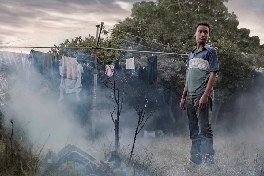 A boy stands in a foggy, overgrown garden beside a Hill Hoist clothesline.