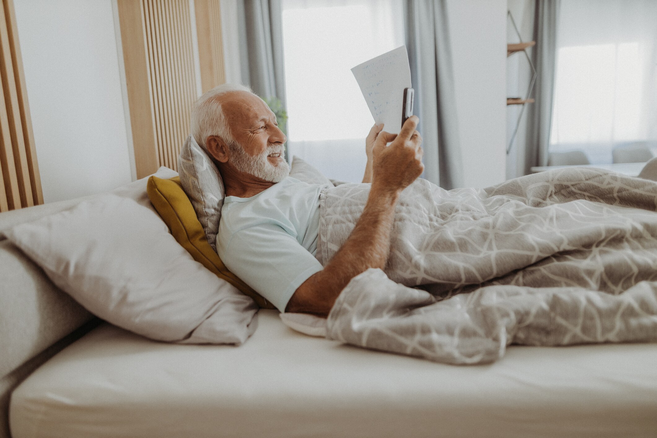An older man with a beard is lying down in bed reading his phone