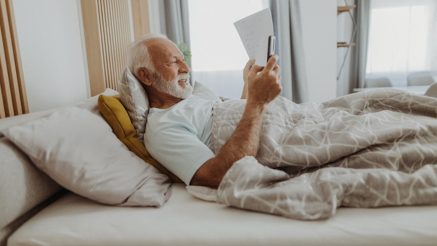 An older man with a beard is lying down in bed reading his phone