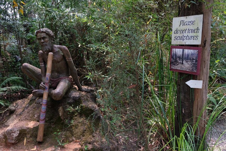 A sculpture of an Aboriginal man with a didgeridoo, next to a sign with a photo of the burnt-out gardens.