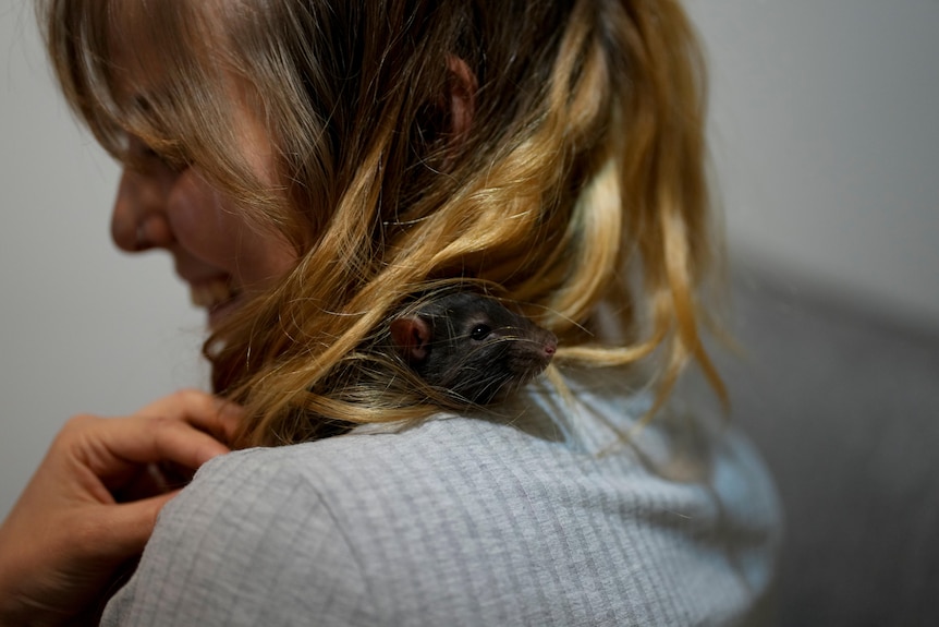 A blonde woman smiles as a dark brown rat rests over her shoulder