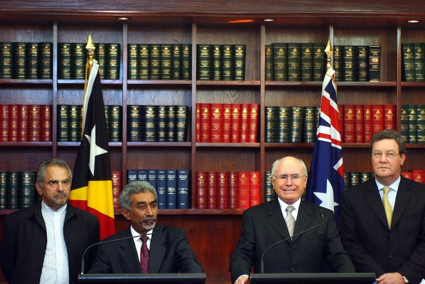 Four men stand behind two lecturns with shelves of books and the Australian and East Timor flags behind them. 