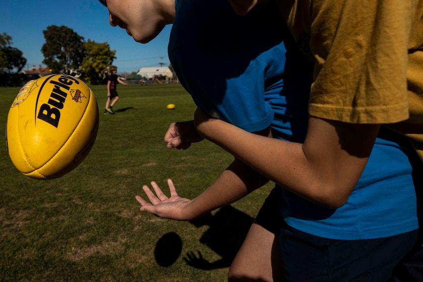 A footballer wearing a yellow shirt tackles a young footballer wearing a blue shirt after he handballs a yellow football.