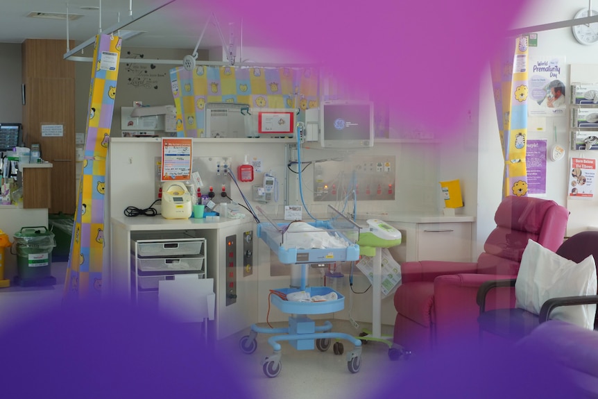 Blurred out pink and purple paper in the foreground, baby, crib, cords and special care unit in background.