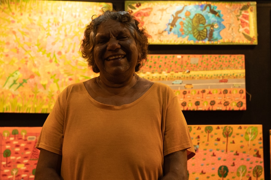 A smiling woman standing in front of a series of brightly colored artworks displayed on a dark wall, inside a gallery.