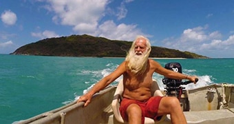 A man with long white hair and beard sails on a tin boat across clear green water, with a small tree-covered island behind him.