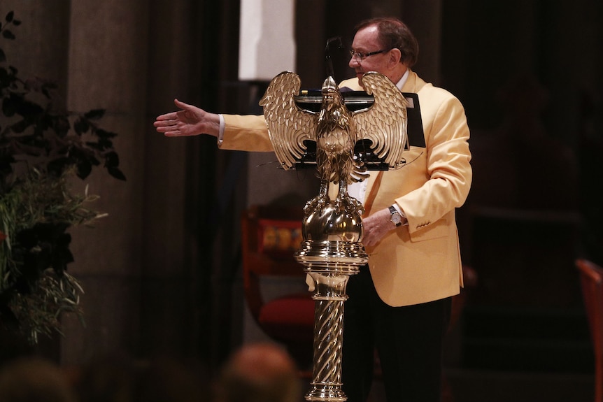 Businessman Harold Mitchell, wearing a bright yellow jacket, stands behind a lecturn and gestures with his hand.