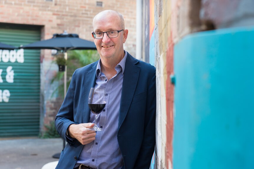 An older, bald man, wearing glasses and dressed in a blue shirt and dark blue blazer, holds a glass of red wine.