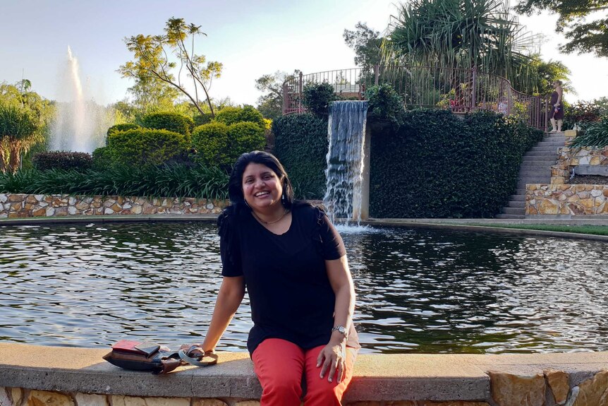A smiling woman sitting in front of a fountain in a tropical environment.