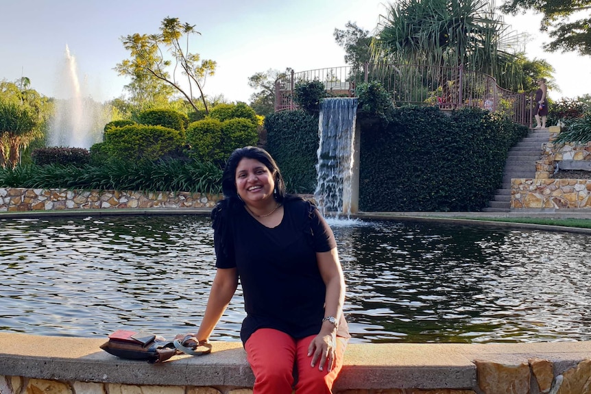 A smiling woman sitting in front of a fountain in a tropical environment.