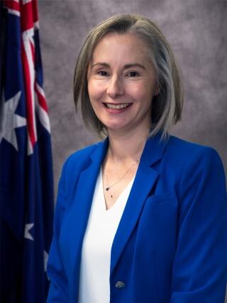 A woman in a blue blazer and shoulder-length hair stands in front of an Australian flag