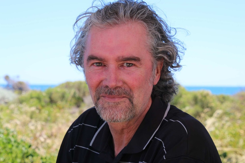 A head and shoulders shot of a man with grey hair and a grey beard with the ocean in the background.
