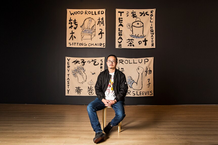 Colour photograph of artist Jason Phu sitting in front of artwork ROLLING ROLLS ROLLED ROLL against a black wall.