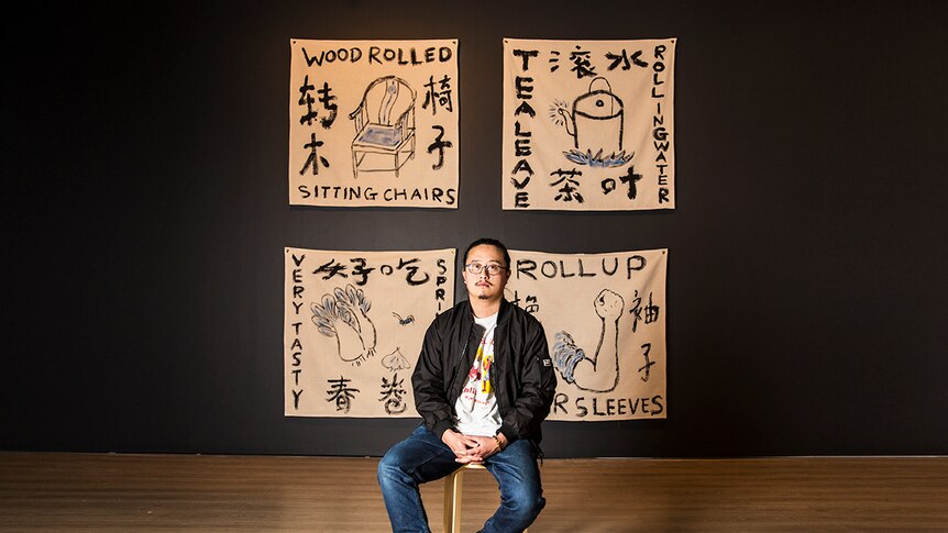 Colour photograph of artist Jason Phu sitting in front of artwork ROLLING ROLLS ROLLED ROLL against a black wall.