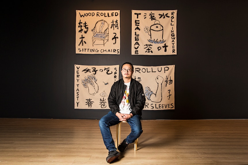 Colour photograph of artist Jason Phu sitting in front of artwork ROLLING ROLLS ROLLED ROLL against a black wall.