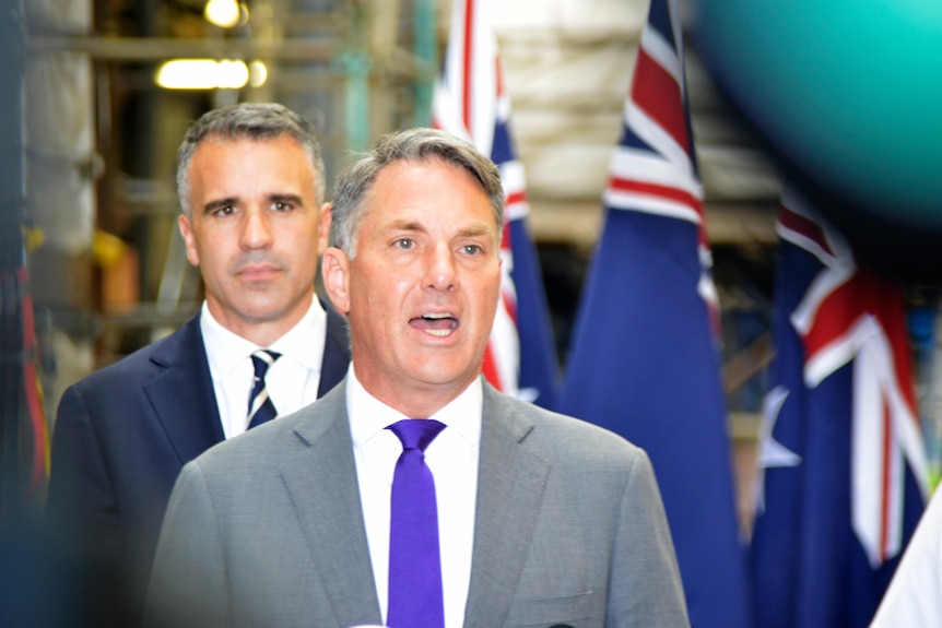 A man stands speaking with another man standing behind him and Australian flags in the background
