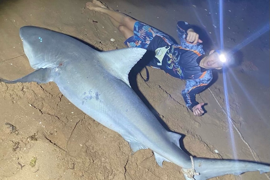 A teenager laying on the sand next to a shark he has caught while fishing.