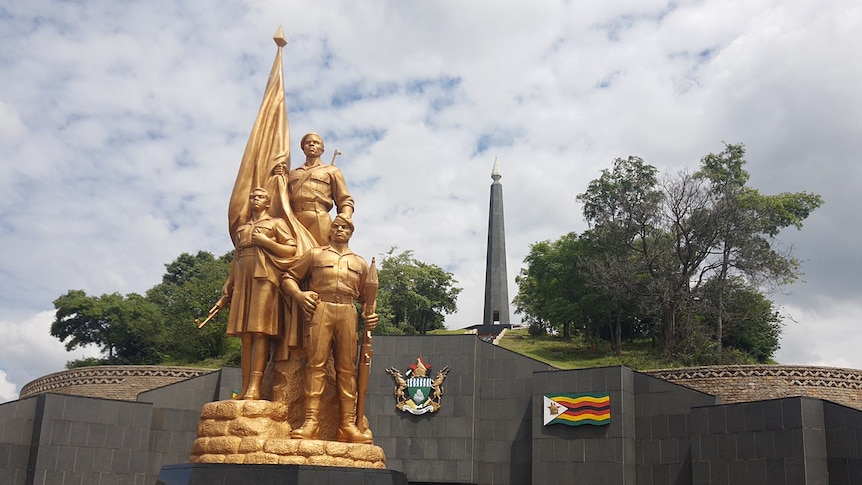 Looking up at a gold statue of male and female Zimbabwean soldiers on a memorial mound made out of dark stone.