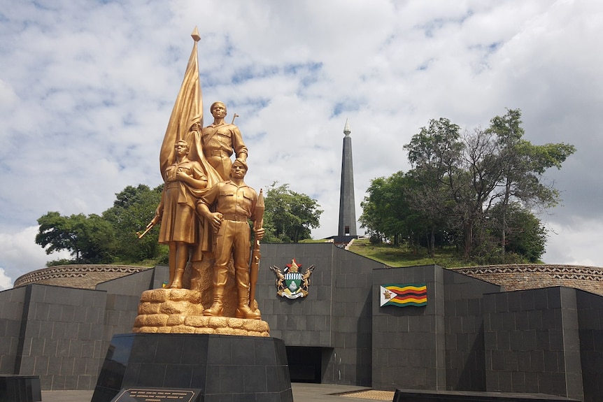 Looking up at a gold statue of male and female Zimbabwean soldiers on a memorial mound made out of dark stone.