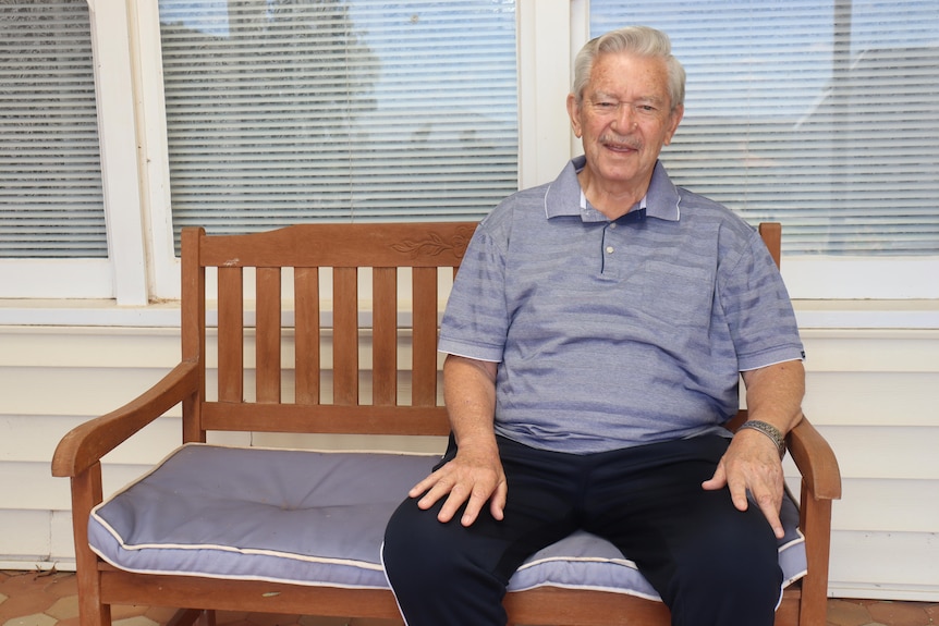 An older man with a blue polo shirt sitting on a wooden bench with a cushion in front of a house.