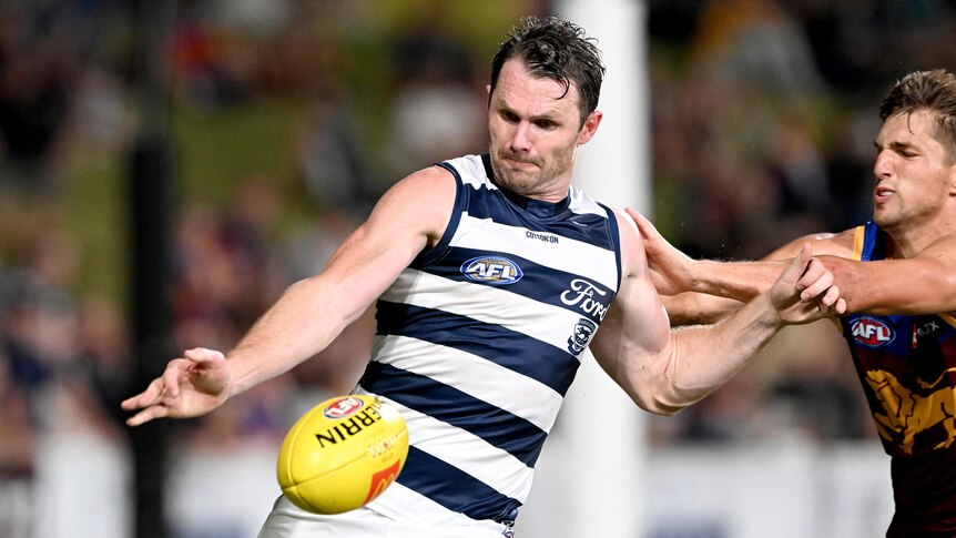 Geelong AFL star Patrick Dangerfield grimaces as he gets ready to kick the ball, while a Brisbane defender tries to grab him.