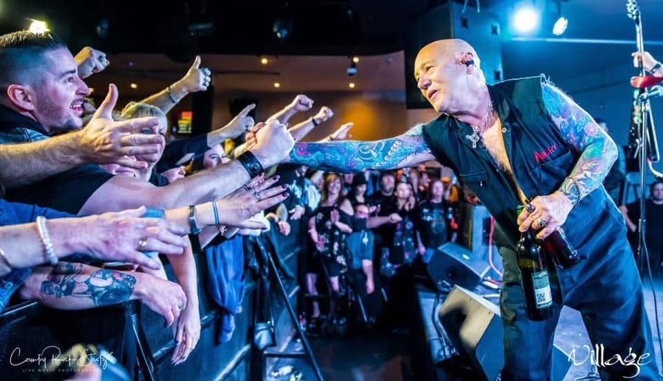 A crowd presses against a barrier reaching out to a bald tattooed man on stage who reaches to them.
