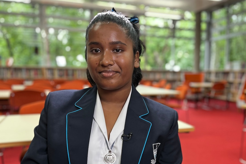 Arlene sits in a classroom. She is wearing a blue blazer and white shirt. There are tables, chairs and red carpet behind her.