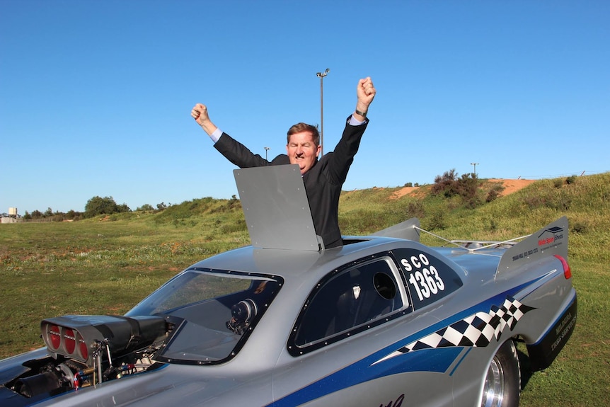 A man standing in the sunroof of a racing vehicle with his hands in the air.