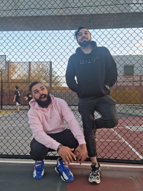 A man kneels next to another man who is leaning on a wire fence, with both wearing tracksuits and smiling.