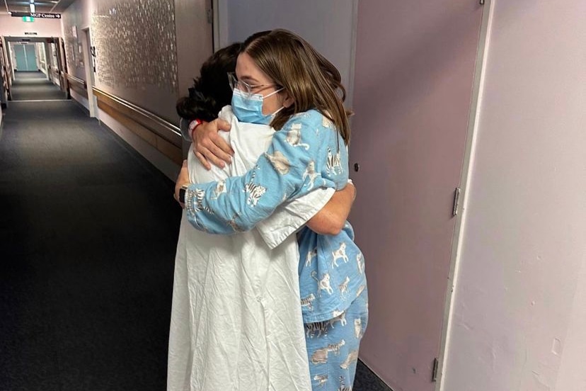 Mother and daughter in hospital robes hug in hospital hallway 
