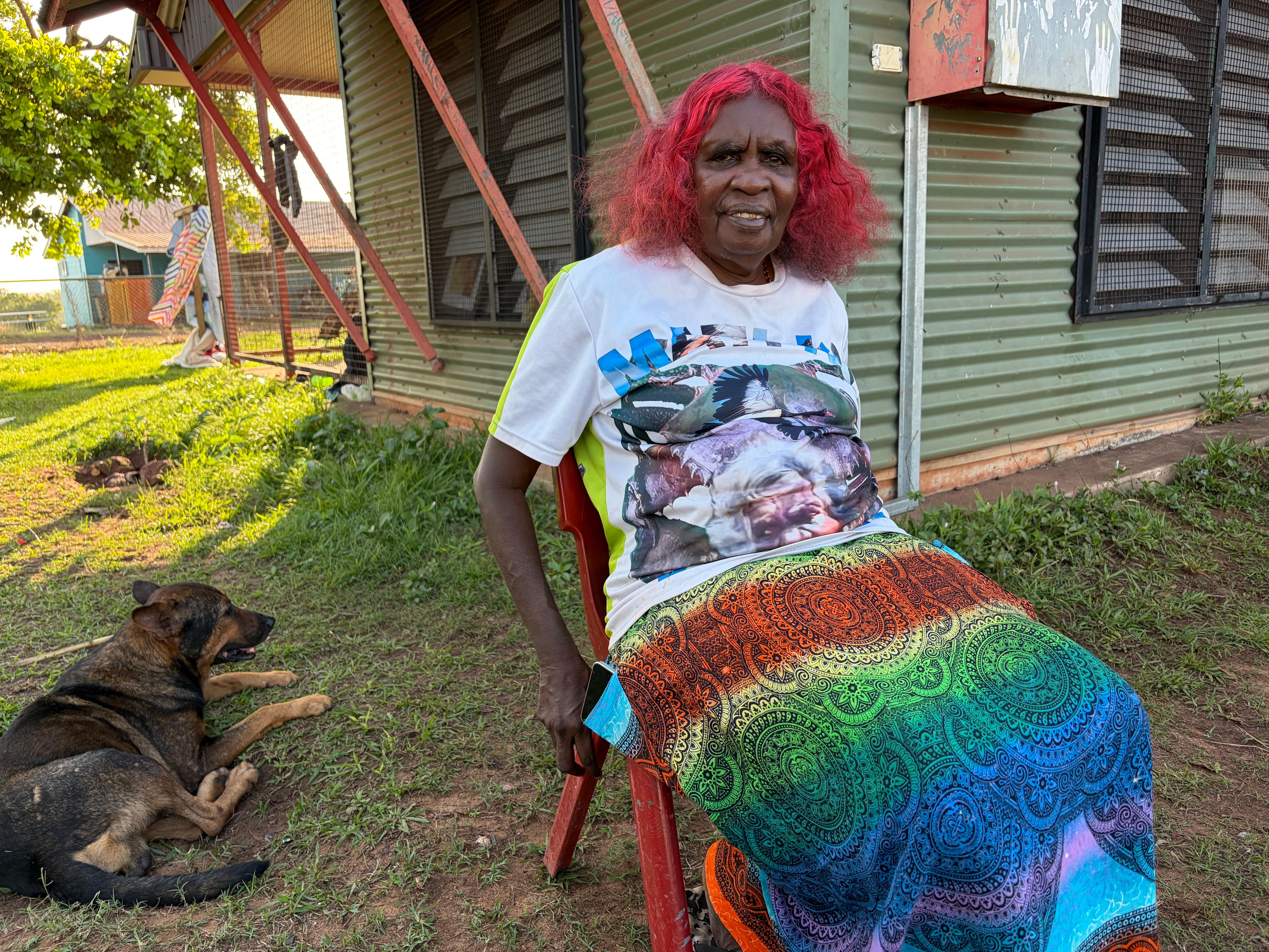 Indigenous woman with red hair dressed in colourful shirt and skirt, Margaret Perdjert, sits down in front of house next to dog.
