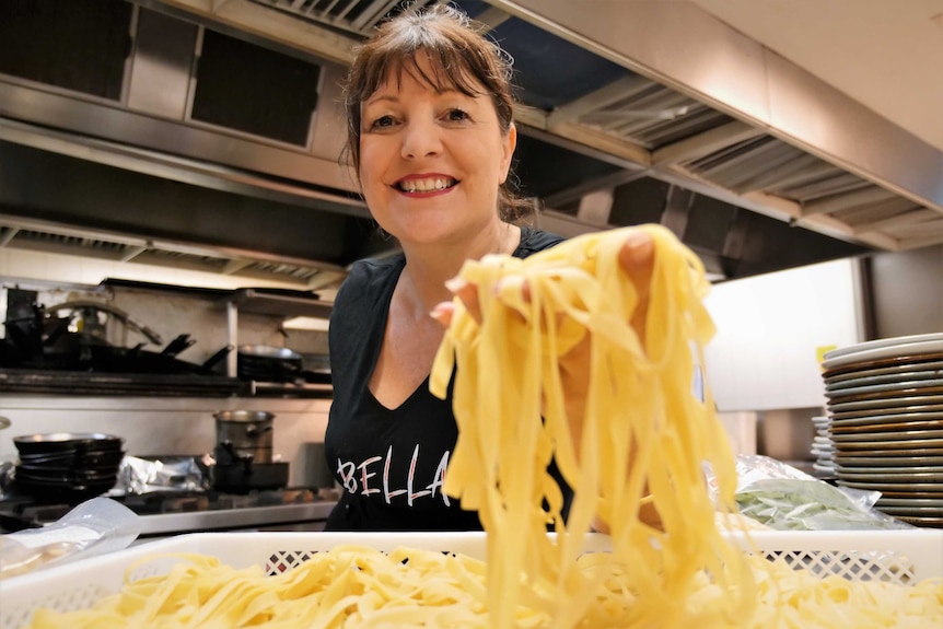 Woman wearing black Bella T-shirt in a commercial kitchen holding up fresh tagliatelle pasta to the lens.